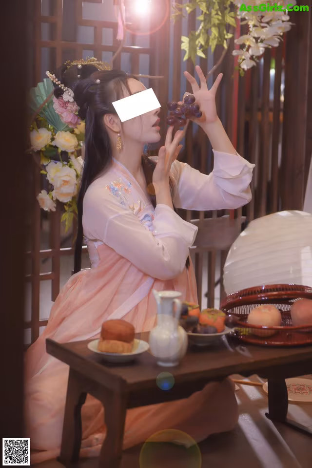 A woman in a pink dress sitting at a table with a basket of fruit.