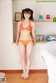 A woman in an orange bikini standing in front of a bed.